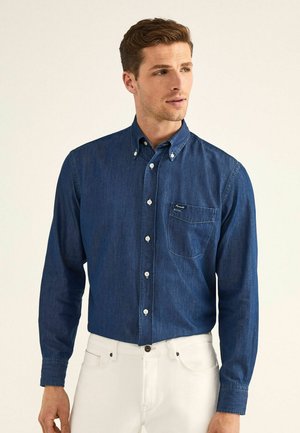 Camisa de mezclilla azul oscuro con mangas largas, cuello abotonado y bolsillo en el pecho. Cuenta con botones blancos y una tela texturizada.