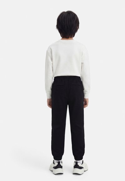 Enfant aux cheveux courts et foncés, vu de dos, portant un haut blanc à manches longues, un pantalon de jogging noir et des baskets blanches, sur fond blanc.