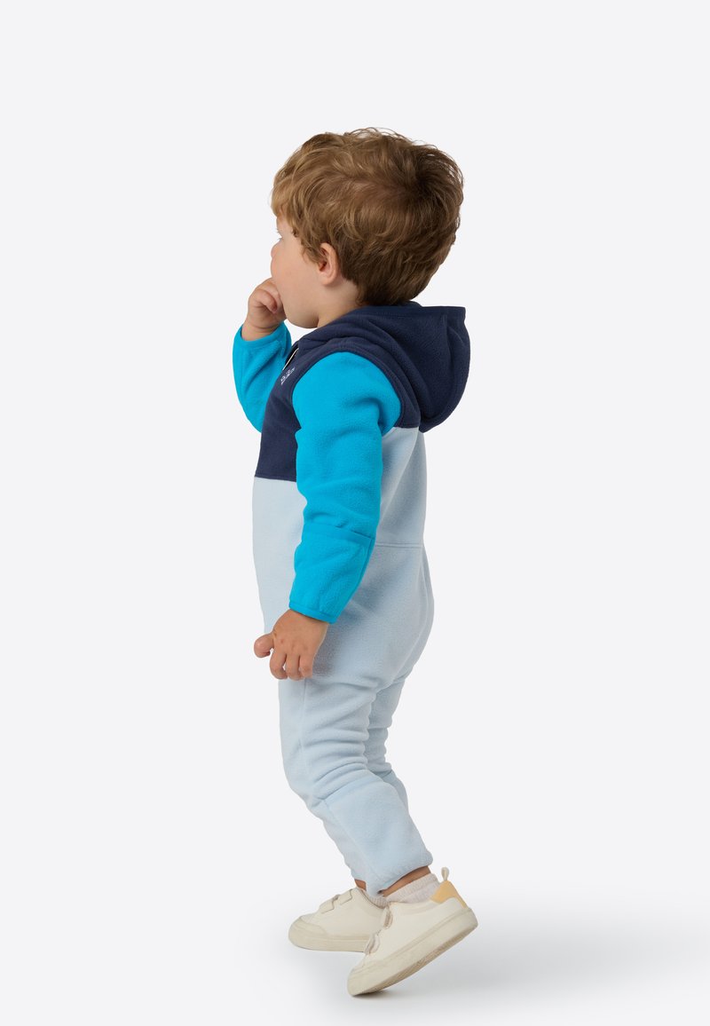 Kombinezon z polaru w jasnoniebieskim kolorze z granatowym kapturem i górną częścią. Charakteryzuje się luźnym krojem, długimi rękawami oraz elastycznymi mankietami. Noszony z jasnymi sneakersami.