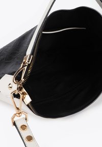 En delvis öppen handväska med en svart insida, gulddragkedja och vit axelrem med ett guldspänne, som visar inga föremål inuti.