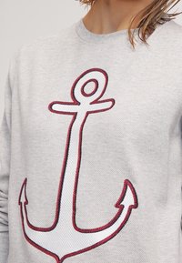 Felpa grigia con un grande design di ancora ricamato in filo bianco e rosso. Il tessuto appare texturizzato e presenta un colletto girocollo.