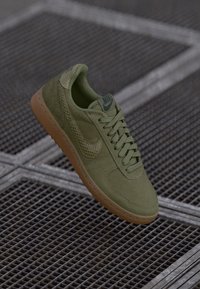 Maslinasto zelena Nike tenisica s teksturiranim swooshom i gumenom potplatkom na pozadini metalne rešetke.