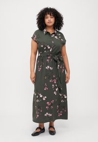 Maxi dress in dark green fabric with short sleeves, floral pink and brown pattern, belted waist, and a collar. Worn with black studded shoes.