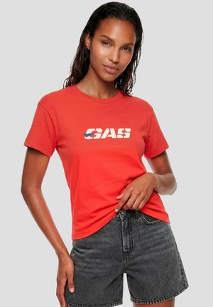 Donna con una maglietta rossa a maniche corte con il logo "GAS" e pantaloncini di jeans grigio scuro, in piedi contro uno sfondo chiaro uniforme.