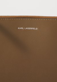 Pochette en faux cuir marron avec fermeture éclair, présentant des coutures visibles et le logo "KARL LAGERFELD" en blanc sur le devant.