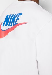 Vit sweatshirt med en framträdande blå och rosa Nike-logotyp med swoosh-design på baksidan, tillverkad av mjukt bomullsmaterial.