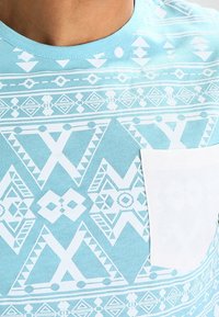 Camiseta turquesa con patrones geométricos blancos, con un escote redondo y un bolsillo blanco en el lado izquierdo del pecho. Fabricada con tela de algodón.