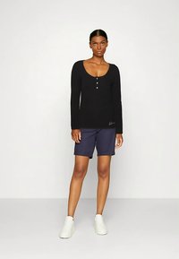 Schwarzes Langarmshirt mit Rundhalsausschnitt und drei Knöpfen, kombiniert mit marineblauen Shorts und weißen Turnschuhen. Schlichtes Design, weicher Stoff.