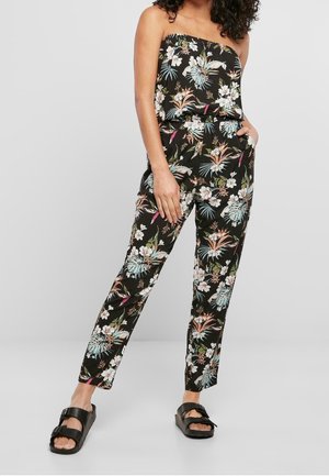 Sort blomsterdragt med en stropløs overdel, der har farverige tropiske mønstre og tætsiddende bukser. Modellen har chunky sorte sandaler på.