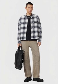 Zwart-wit geruit overhemd met vredessymboolpatronen, gecombineerd met een zwart t-shirt en beige broek. Draagt een zwarte tas en zwarte schoenen.