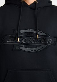 Μαύρη φούτερ με καπάκι που διαθέτει μια υφασμάτινη, πλαστική ανάγλυφη λογότυπο Dickies σε μαύρο χρώμα. Κορδόνια με χρυσές μεταλλικές άκρες, τσέπη καγκουρό μπροστά.