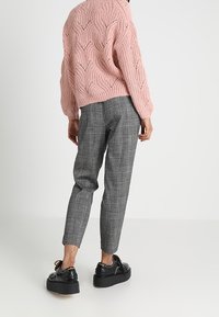 Kobieta stoi, ubrana w różowy sweter z dzianiny, szare dopasowane spodnie w kratę i czarne buty na platformie na jednolitym białym tle.