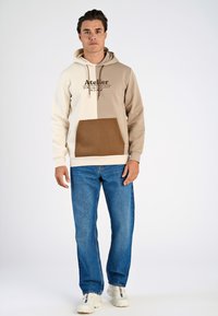 Hoodie in beige en lichtbruin, met een bruine zak, bedrukte logo op de borst, gecombineerd met blauwe denim jeans en sneakers.