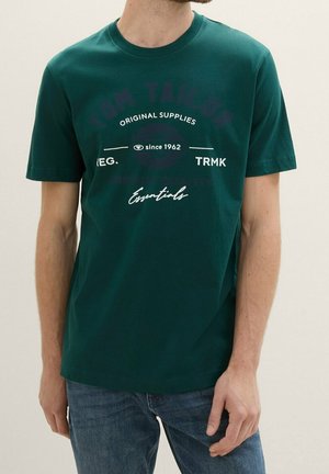 Mężczyzna w ciemnozielonym t-shircie z białym napisem „Original Supplies since 1962 REG. TRMK Essentials” i niebieskich dżinsach na jednolitym tle.