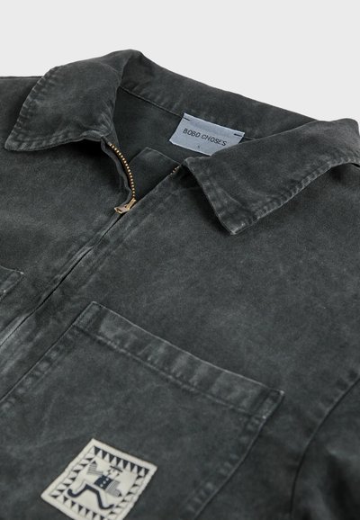 Dunkelgraues, strukturiertes Kurzarmhemd mit einem Reißverschlusskragen, das mit einem wei�ßen Patch mit geometrischem Design und zwei frontalen Taschen ausgestattet ist.