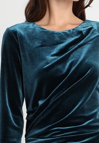 Teal sammetstopp med båtringning, långa ärmar och draperad detalj över framsidan, som visar en slät, glänsande yta.