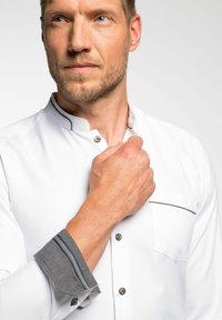 Weißes Hemd mit Stehkragen, ausgestattet mit grauen Akzenten an den Manschetten und einer Brusttasche. Weiches Material mit glatter Textur.