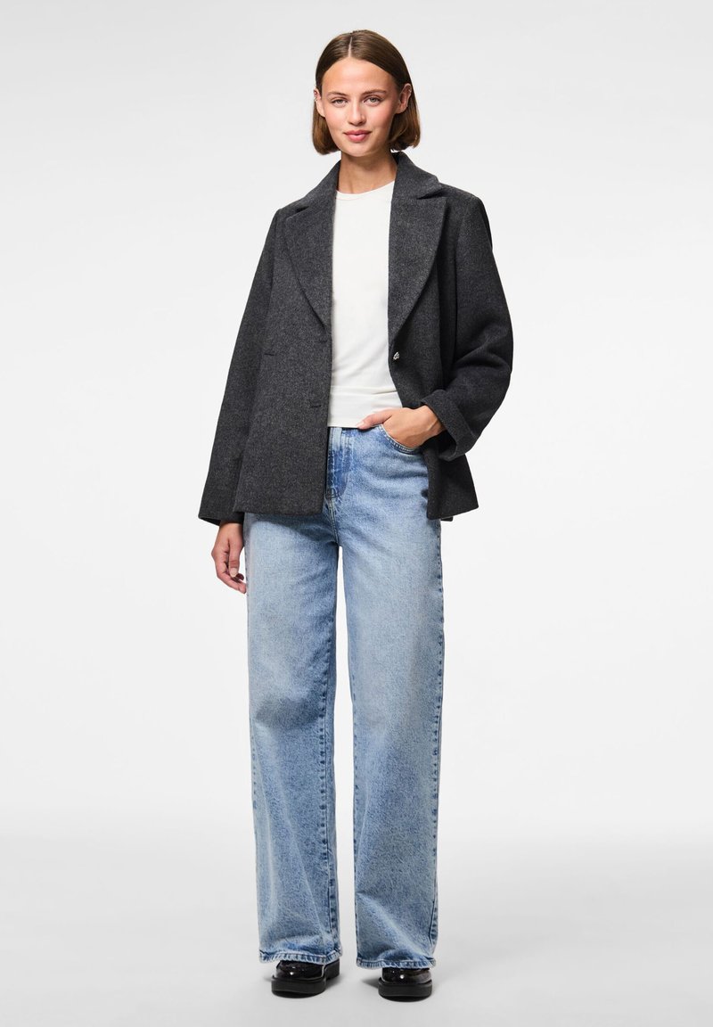 Grå ullblandad jacka med slagkrage, dubbelknapplockning och avslappnad passform, kombinerad med ljusblå vida jeans och svarta skor.