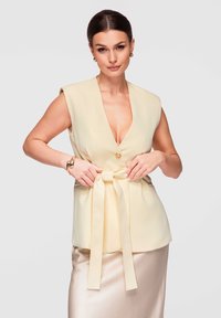 Femme aux cheveux foncés portant un gilet sans manches jaune pâle noué à la taille et une jupe brillante beige clair, sur un fond uni.