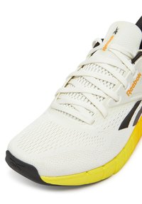 Białe buty sportowe z siateczkową cholewką, czarnymi akcentami, logo Reebok i żółtą podeszwą. Posiadają zaokrąglony czubek oraz fakturowane detale na powierzchni.