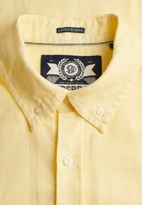 Col de chemise en mélange de lin jaune avec des boutons blancs et une étiquette noire portant la mention "Linen Blend" ainsi qu'une étiquette de marque à l'intérieur du col.