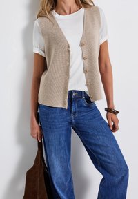 Kobieta ubrana w beżową, zapinaną na guziki dzianinową kamizelkę na białą koszulkę z podwiniętymi rękawami i niebieskie dżinsy, trzyma brązową zamszową torbę i nosi ciemną bransoletkę.