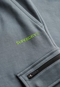 Szara bluza z gładkiego materiału, z zielonym logo "SUPERDRY" i detalem kieszeni na zamek.