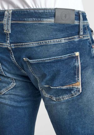 Jean en denim bleu avec un design classique, cinq poches et une finition texturée, avec une étiquette de ceinture en cuir gris et des coutures contrastées.