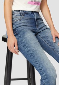 Jasnoniebieskie dżinsy o dopasowanym kroju, z przednimi kieszeniami i delikatnym przecieraniem. Na udzie spoczywa ręka, siedząca na stołku.