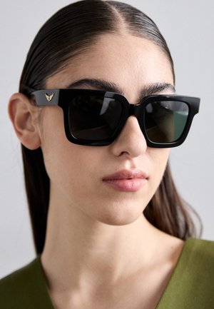 Black square sunglasses with dark lenses, featuring a gold wing detail on the temple. Matte finish and bold, angular design.
