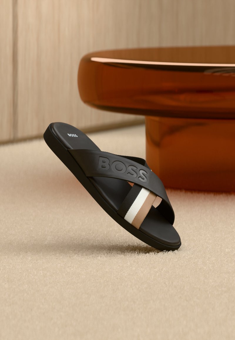 Μαύρη σαγιονάρα BOSS με ανάγλυφο λογότυπο και λεπτομέρεια από ύφασμα με ρίγες σε καφέ, λευκό και μπεζ, σε μοκέτα δίπλα σε καφέ βάση τραπεζιού.