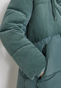 Pufi bunda v tlumené tyrkysové barvě, s vrchní částí z manšestru s ribbenou texturou a hladkým dolním dílem. Obsahuje detail se zipovou kapsou.