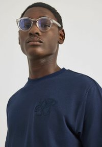 Granatowa bawełniana koszulka z krótkim rękawem, z teksturowanym haftowanym logo i okrągłym dekoltem. Przezroczyste okrągłe okulary z niebieskimi szkłami.