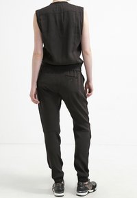 Czarny kombinezon bez rękawów z dopasowanym wcięciem w talii, bocznymi kieszeniami i zwężanymi nogawkami; gładki materiał z minimalną ilością szwów; noszony z wzorzystymi sneakersami.