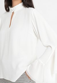 Blusa bianca con colletto alto, scollo a chiave e maniche a trombetta. Tessuto liscio, leggero e con una lieve lucentezza. Abbinata a pantaloni a scacchi.