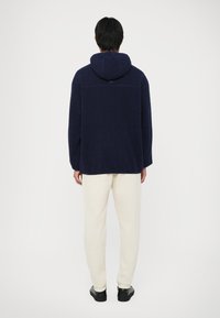 Sweatshirt de fleece azul marinho com um acabamento texturizado, um capuz grande e um corte descontraído, combinado com calças off-white e sapatos pretos.