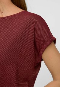 Nærbillede af en person iført en kortærmet, maroon skjorte med tynde horisontale hvide striber og en opkrøllet ærmekant.