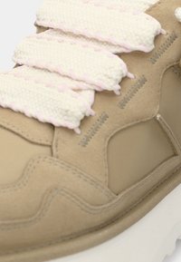 Μπεζ σουέτ αθλητικό παπούτσι με υφασμένα κρεμ κορδόνια που έχουν ροζ λεπτομέρειες, λεία, στρογγυλεμένη μορφή και παχιά λευκή σόλα.