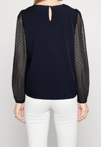 Blouse bleu marine avec manches transparentes à pois, détail dos avec ouverture en trou de serrure et coupe décontractée. Associée à un pantalon blanc ajusté.