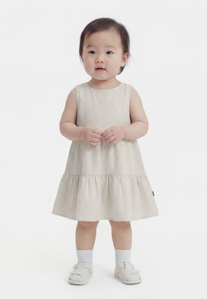 Småbarn stående iført en ærmeløs beige stribet kjole, hvide sokker og hvide sandaler med hænderne foldet foran.