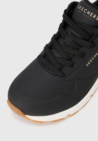 Black sneaker with perforated upper, round toe, and flat laces. Features a white midsole and a gum rubber outsole. Brand name visible on the side.