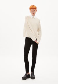 Cremfarbener Strickpullover mit rundem Ausschnitt, kombiniert mit schwarzen, enganliegenden Hosen und schwarzen Clogs. Verfügt über lange Ärmel und eine strukturierte Oberfläche.