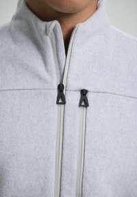 Svetle šedá fleecová bunda s vysokým límcem, se zipovým uzávěrem a černými táhly zipu. Měkká textura bez viditelných vzorů nebo akcentů.