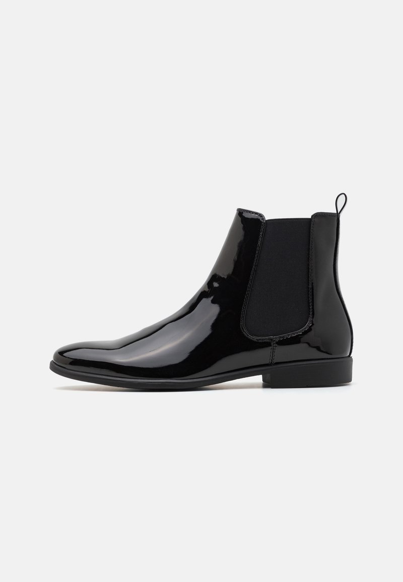 Bottine Chelsea noire brillante avec panneau latéral élastique, languette au dos et petit talon plat sur fond blanc uni.