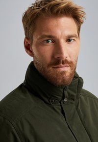 Tmavě zelená bunda s vysokým límcem, zapínáním na patentky a texturovanou tkaninou. Disponuje jemným leskem a střihem, který zvýrazňuje postavu.