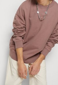 Sudadera marrón de tela mixta con cuello redondo, corte oversize y puños de canalé, combinada con pantalones de color claro y un collar de cadena de plata.