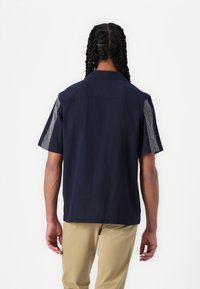 Camisa de manga corta de tela azul marino, con acentos con estampados en los hombros. Cuello clásico y corte relajado. Textura suave.