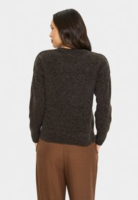 Mujer con cabello largo y oscuro, vestida con un suéter texturizado marrón oscuro y pantalones marrones, de pie de espaldas con las manos en los bolsillos.