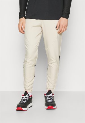 Person iført beige bukser med sorte accenter og sorte sneakers med hvide snørebånd og røde såler, stående mod en ensfarvet baggrund.