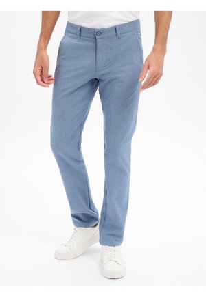 Man draagt lichtblauwe slim-fit broek en witte sneakers, staand tegen een witte achtergrond.